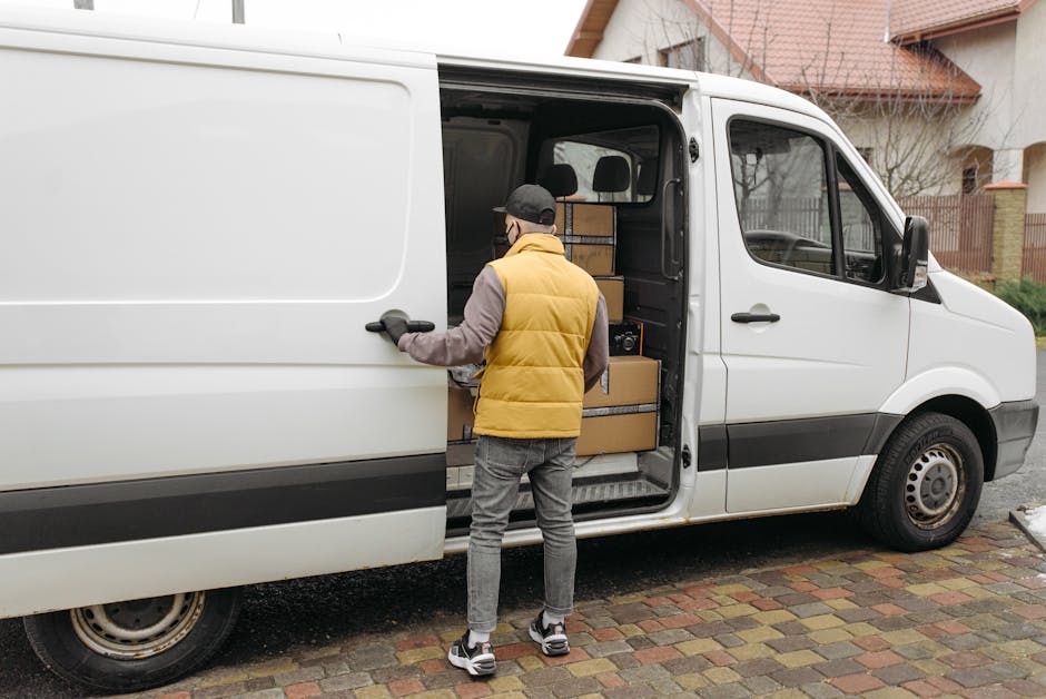 A man in a yellow padded vest, grey hoodie, grey jeans, and sneakers is loading several cardboard boxes into the back of a white panel van with sliding doors, parked on a paved driveway outside residential houses. The boxes are of various sizes and are packed neatly inside the van, some secured with straps for transportation. The background features a brown brick house with a pitched roof, a small tree, and a decorative fence on the right side. The scene depicts the process of home relocation or furniture transport, with the man assisting in packing or moving items during a house removal service. The van's interior is visible, showing more boxes and packing materials, consistent with professional removal logistics provided by Man and Van Lisson Grove, supporting efficient packing and loading during house removals or moving services in the London NW1 area.