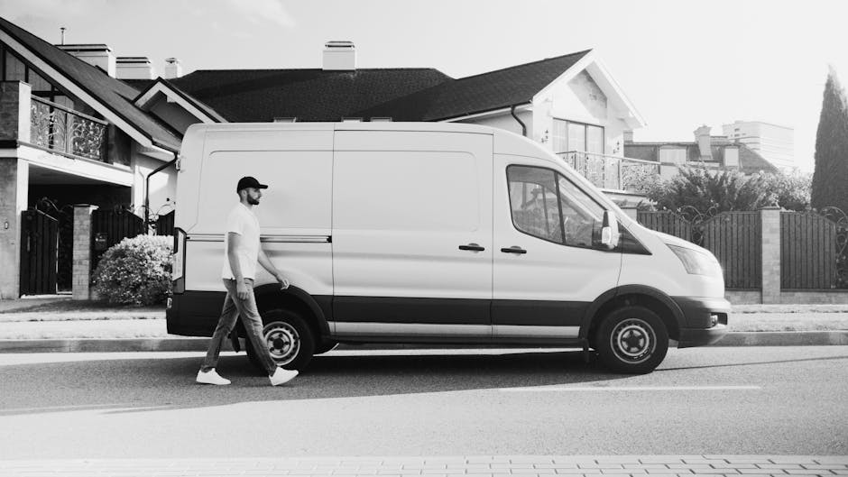 A man wearing a white t-shirt, dark trousers, and a cap is walking alongside a white cargo van parked on the street in a residential neighbourhood. The van is a commercial vehicle used for home relocation and furniture transport, with sliding side doors and a black bumper. Behind the vehicle, there are modern houses with pitched roofs, balconies, and gardens enclosed by fences, suggesting a suburban area in Lisson Grove. The man appears to be preparing for a loading or unloading process, characteristic of packing and moving activities. The scene is captured during daylight with natural lighting, and the van is positioned parallel to the pavement, indicating a typical loading area for house removals. Man and Van Lisson Grove's service involves these types of vehicle-based moving logistics, emphasizing careful handling of furniture, boxes, and household appliances during home relocations.