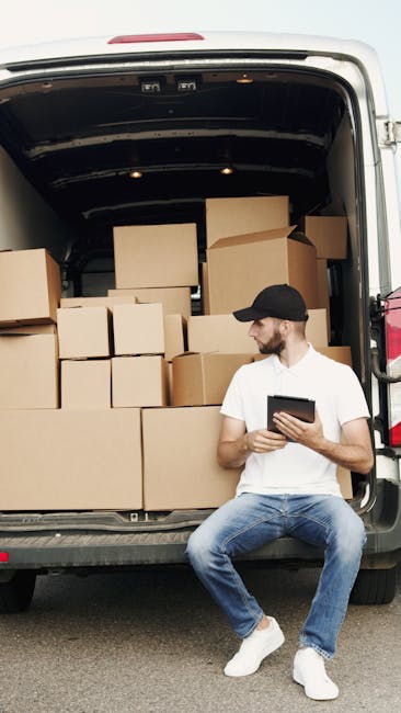 A man in a yellow padded vest, grey hoodie, grey jeans, and sneakers is loading several cardboard boxes into the back of a white panel van with sliding doors, parked on a paved driveway outside residential houses. The boxes are of various sizes and are packed neatly inside the van, some secured with straps for transportation. The background features a brown brick house with a pitched roof, a small tree, and a decorative fence on the right side. The scene depicts the process of home relocation or furniture transport, with the man assisting in packing or moving items during a house removal service. The van's interior is visible, showing more boxes and packing materials, consistent with professional removal logistics provided by Man and Van Lisson Grove, supporting efficient packing and loading during house removals or moving services in the London NW1 area.