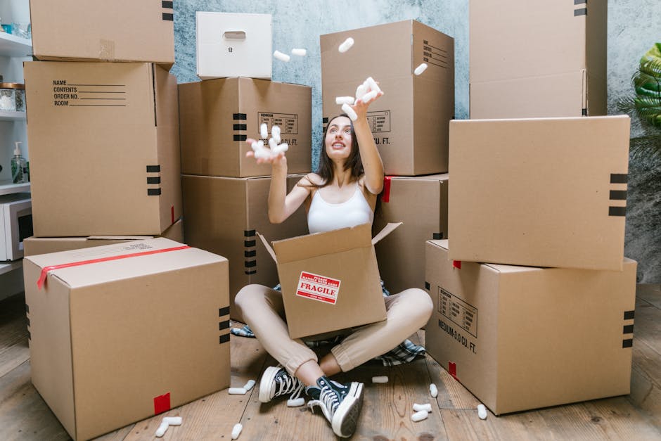 Two removals workers from [COMPANY_NAME] are inside a well-lit room with white walls and large arched windows, preparing for a house relocation. One man, with a beard and curly hair, is carrying a med