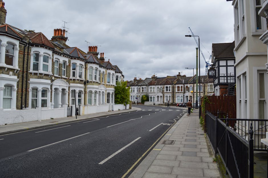 A quiet residential street lined with Victorian-style terraced houses featuring white facades, large bay windows, and tiled roofs topped with red chimney pots. The pavement runs alongside the row of properties, with modern street lighting and a few trees visible further down the street. A man is seen walking away in the distance, while parked cars occupy spaces along the curb. The overcast sky above suggests a cloudy day. This scene is representative of a typical London neighbourhood suitable for house removals and furniture transport in a professional home relocation process. Man and Van Lisson Grove operates in this area, providing expert moving and packing services to facilitate efficient house removals along Marylebone Road and surrounding streets.