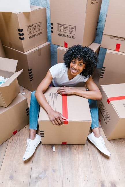 A person wearing light clothing is inside a room with wooden flooring, engaged in packing or preparing for a home relocation by handling a cardboard box. The individual is gripping a medium-sized box 